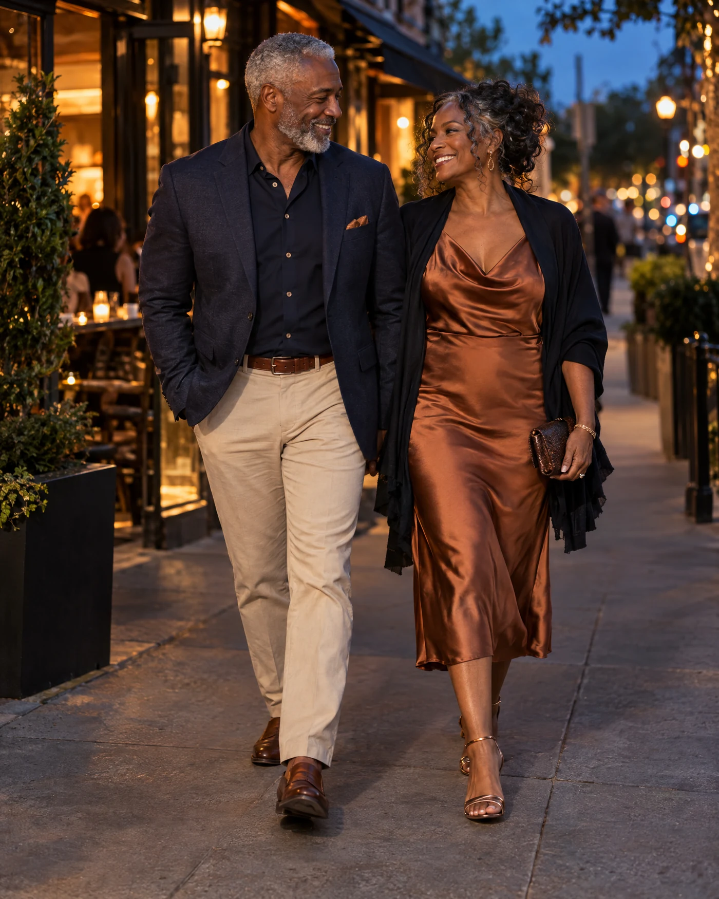A stylish mature couple smiling at each other while walking out together at dusk, the gentleman with short silver hair and beard